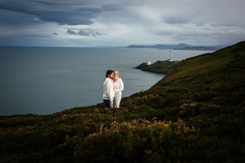 Two people stand close together on a grassy hillside overlooking the Irish sea, with Bailey lighthouse on a distant headland and mountains in the background under a cloudy sky following their secret proposal howth cliffs dublin.