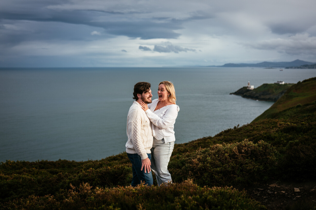 A man and a woman stand together on a grassy hillside overlooking the sea, with cloudy skies and distant cliffs in the background following their secret proposal howth cliffs dublin.