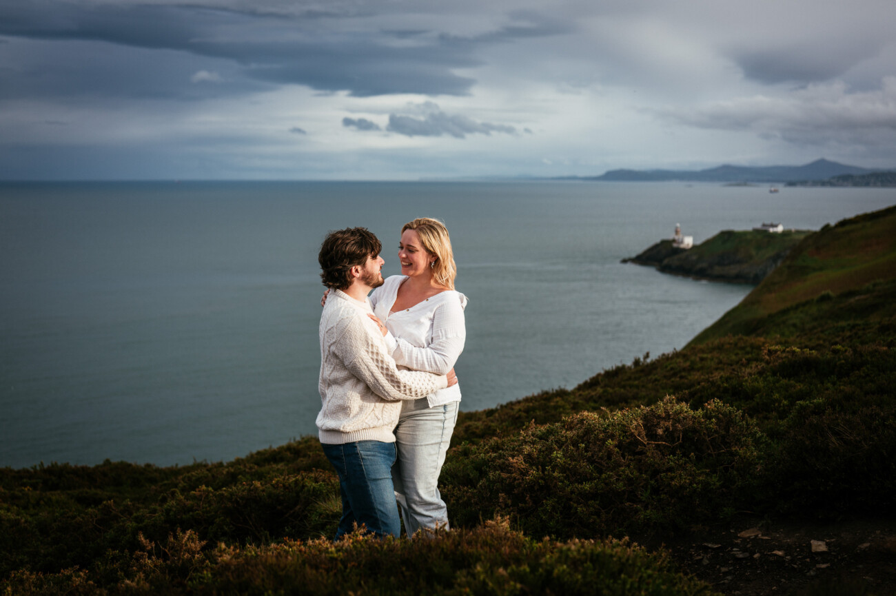 A couple stands on a grassy hill overlooking the sea, with cloudy skies and distant cliffs in the background.