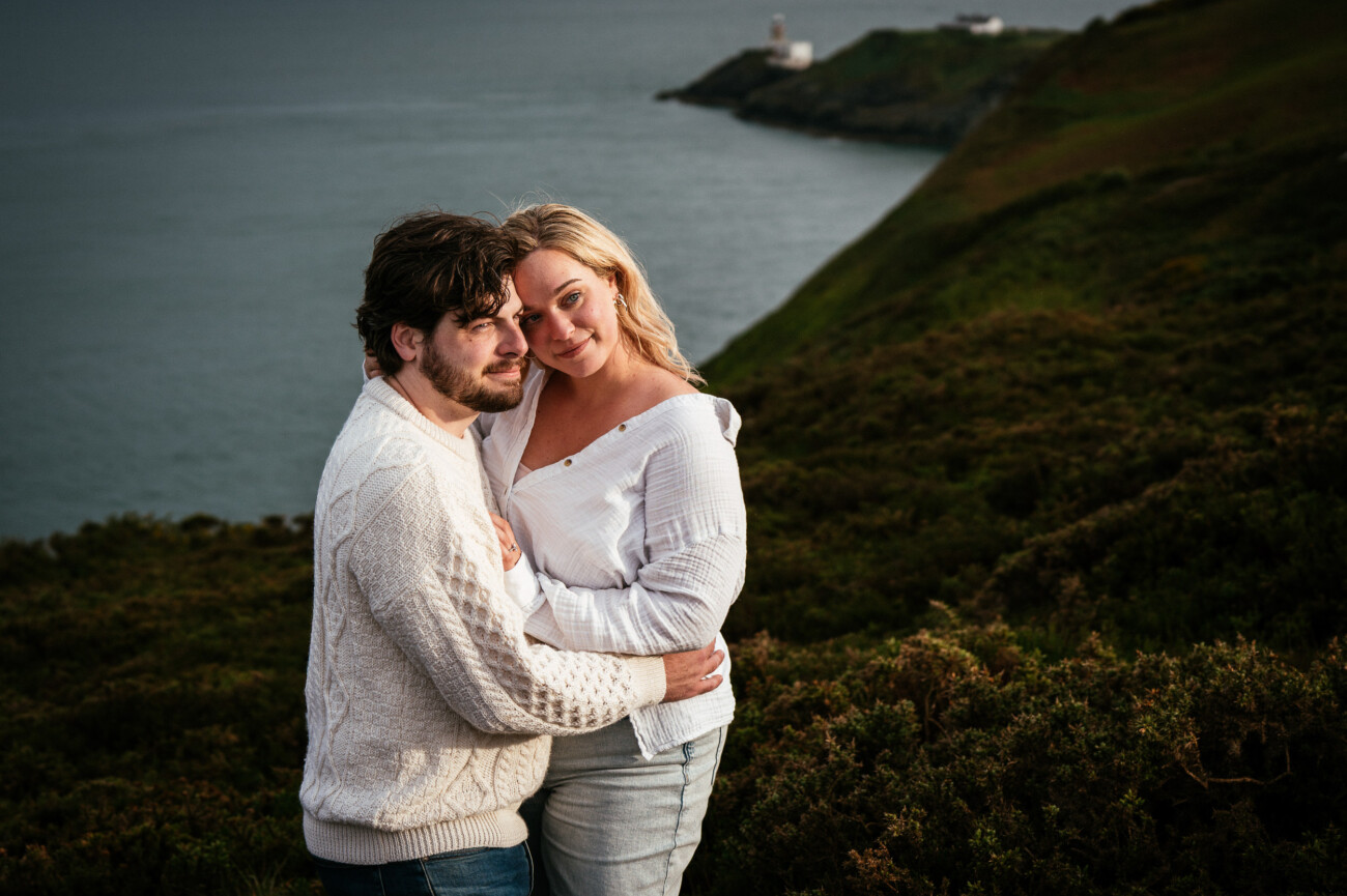 A couple stands close together on a grassy hillside overlooking the sea, with a distant lighthouse visible on a headland in the background ollowing their secret proposal howth cliffs dublin.