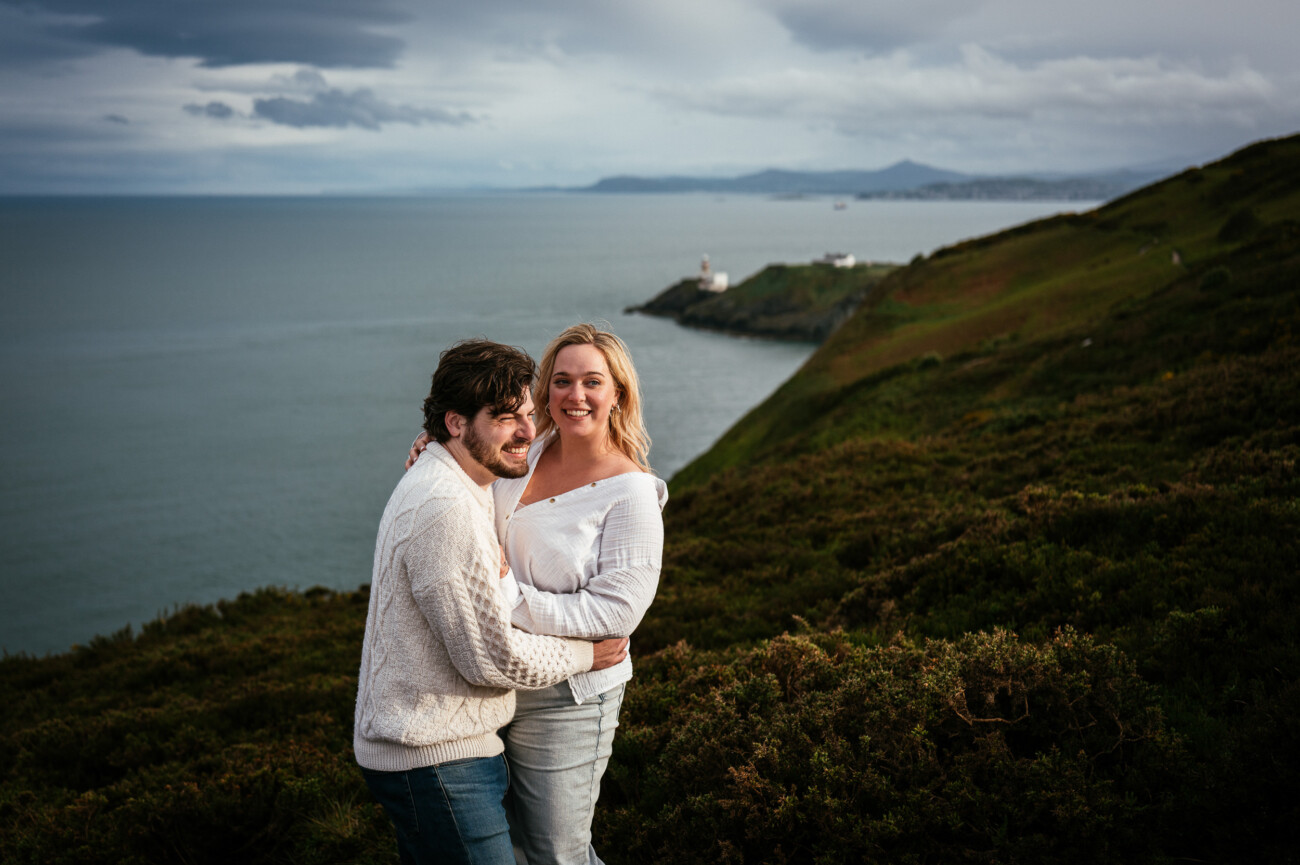 Two people stand close together and smile, with coastal cliffs, green hills, and the sea in the background under a cloudy sky.