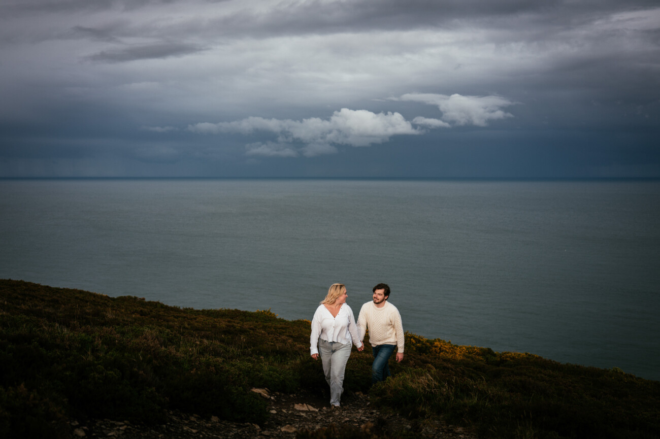 Two people walk hand in hand up a grassy hill overlooking a calm sea under a cloudy sky.