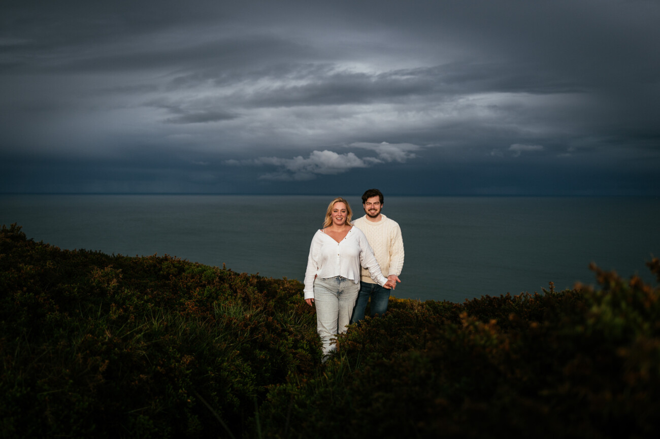 A couple stands together on a grassy cliff overlooking the sea, with dark clouds and a moody sky in the background.