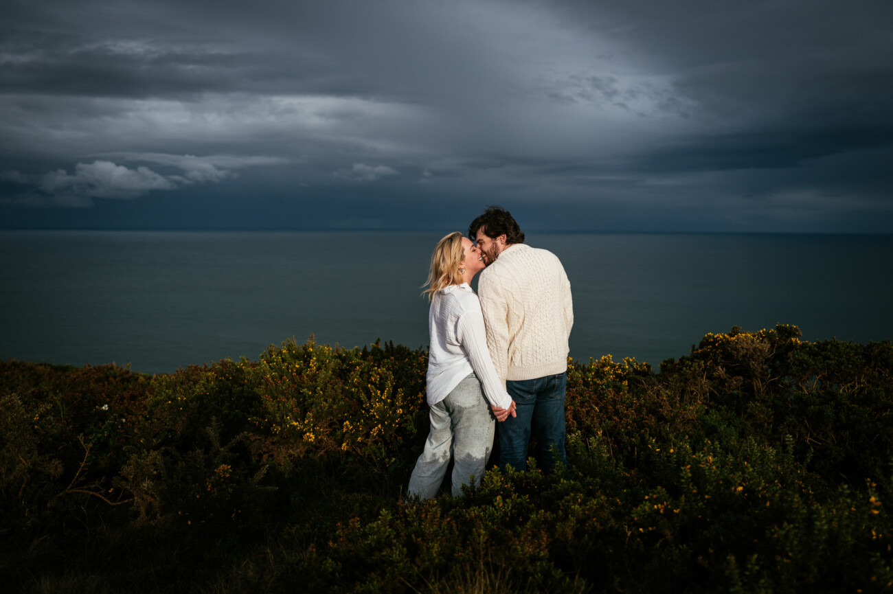 A couple stands close together on a grassy cliffside overlooking the sea under a cloudy sky.