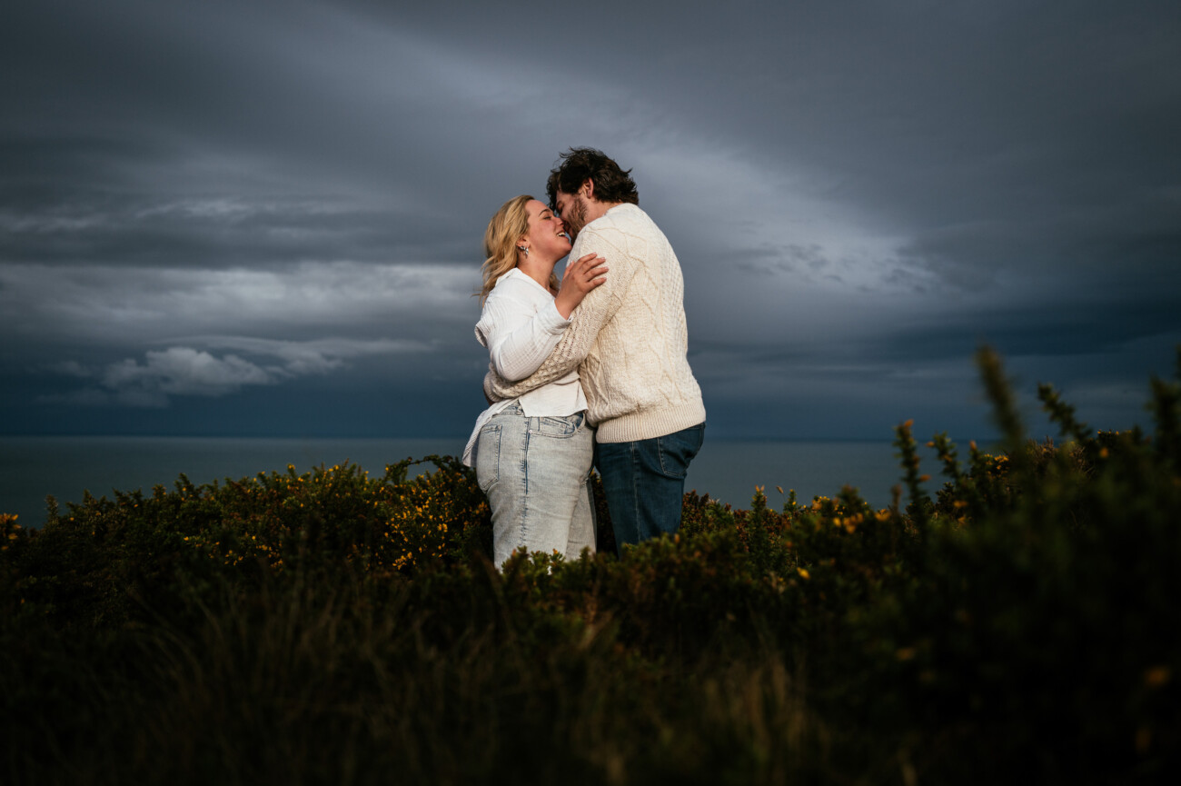 A couple stands closely together, smiling and embracing, on a grassy area with dark, cloudy skies and the sea in the background ollowing their secret proposal howth cliffs dublin.
