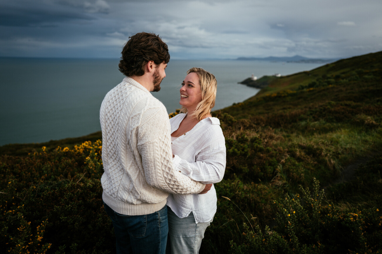 A couple stands close together on a grassy hill overlooking the sea, with cloudy skies and distant cliffs in the background.