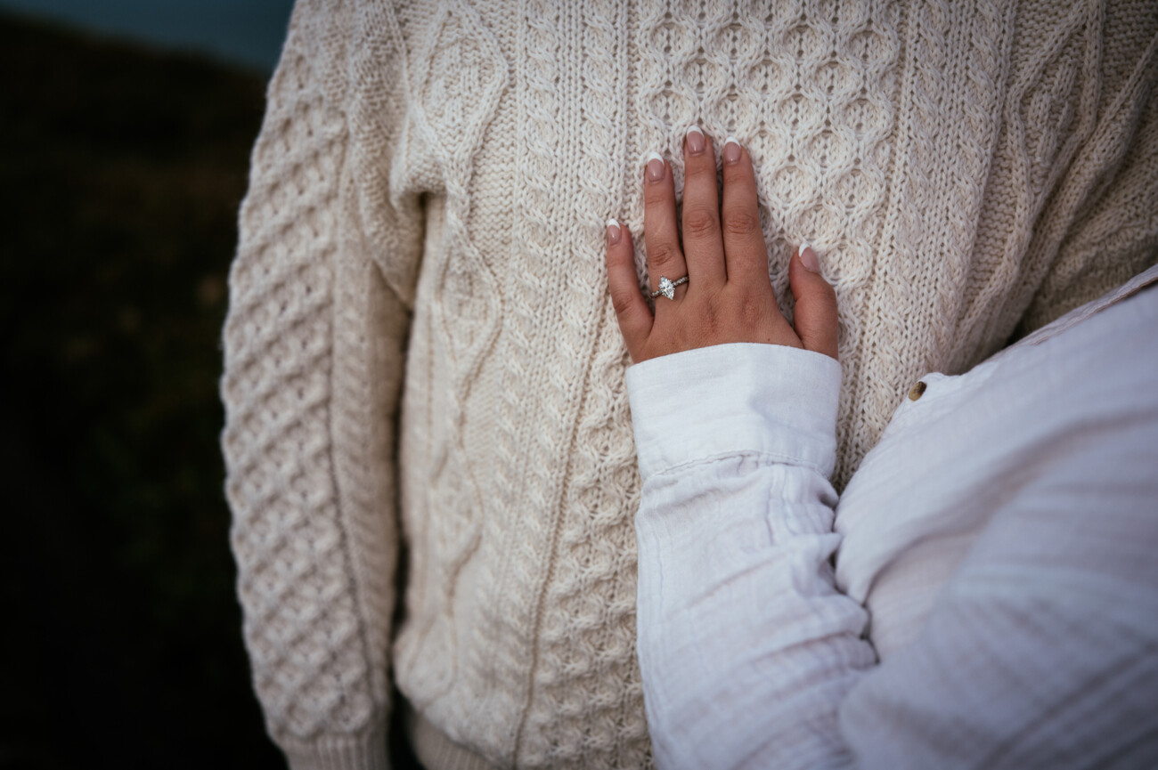 A person wearing a white shirt rests their hand, showing an engagement ring, on the chest of someone in a cream cable-knit jumper.