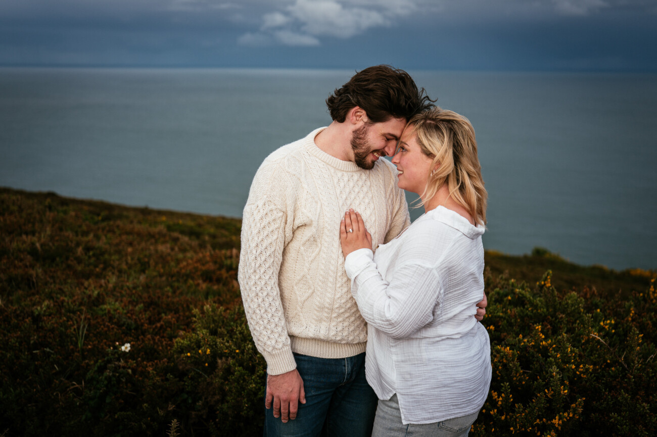 A man and woman stand close together on a grassy cliffside with the sea in the background under a cloudy sky.