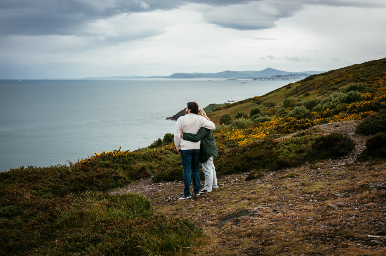 Two people stand on a coastal hillside path, embracing and facing the sea under a cloudy sky, with green shrubs and distant shoreline in the background at Howth Summit, Dublin.
