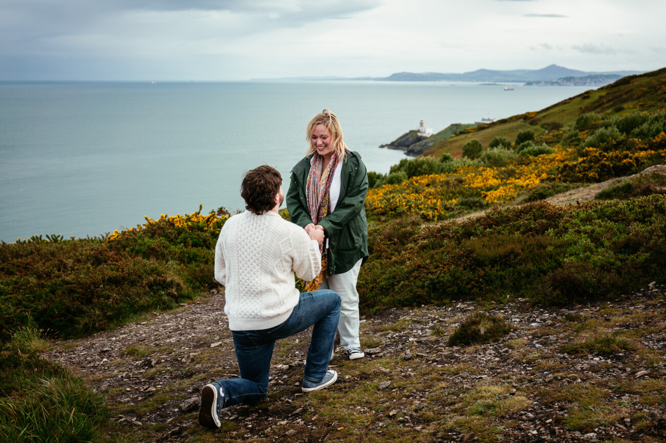 A person kneels and proposes to another person on a coastal hillside, with the sea and distant cliffs visible in the background.