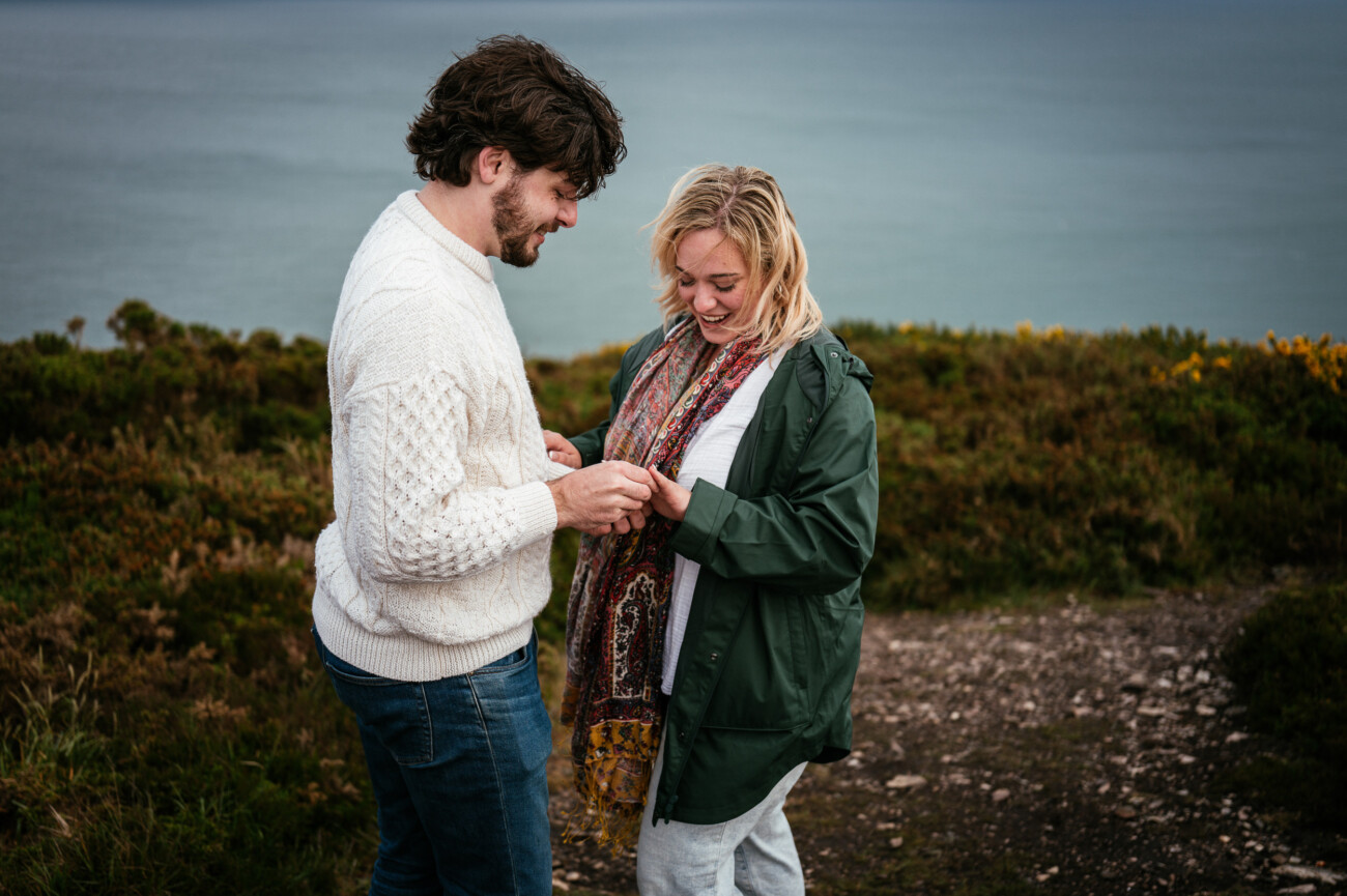 A man and woman stand outdoors on a grassy hill by the water, smiling as the man hands something small to the woman.