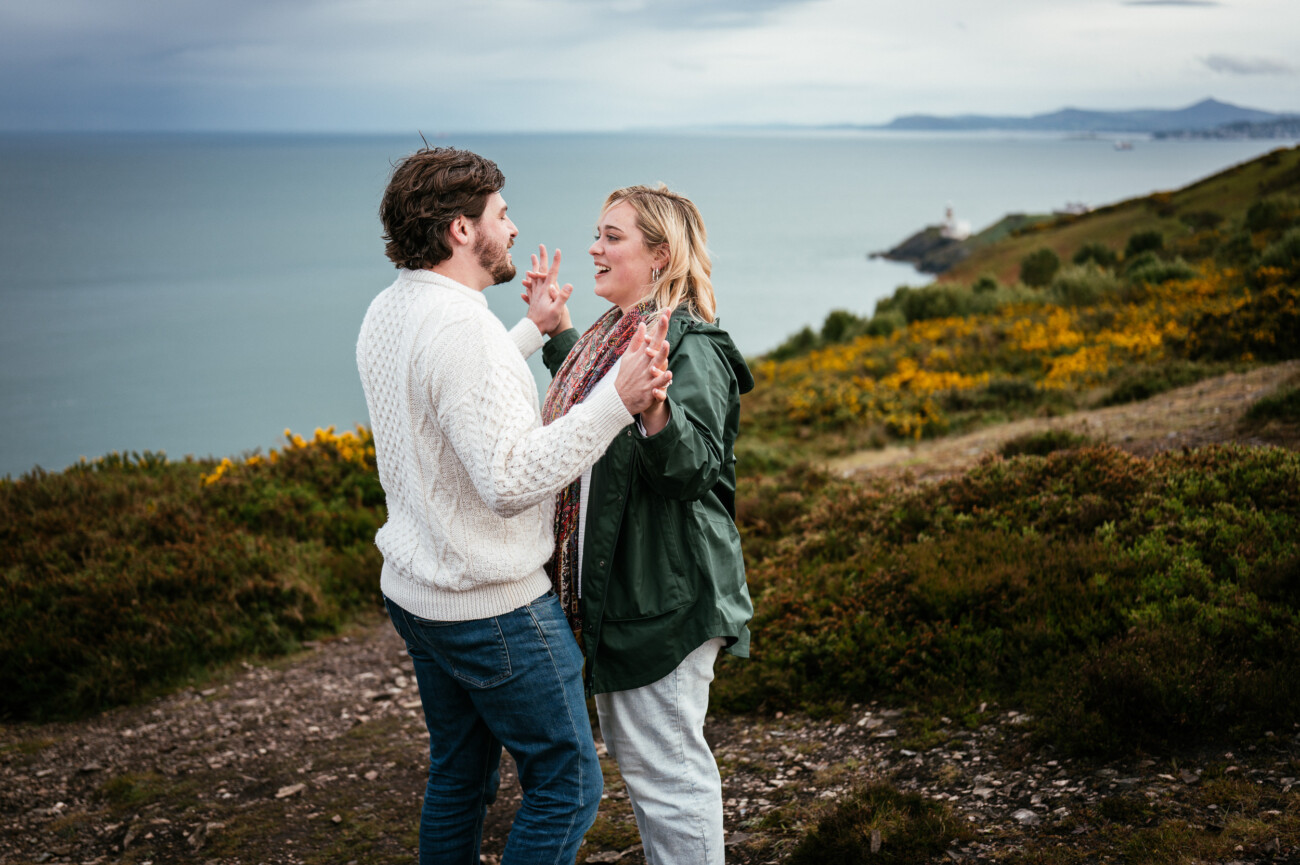 A man and woman stand facing each other on a coastal path, gesturing whilst talking, with the sea and hills in the background.
