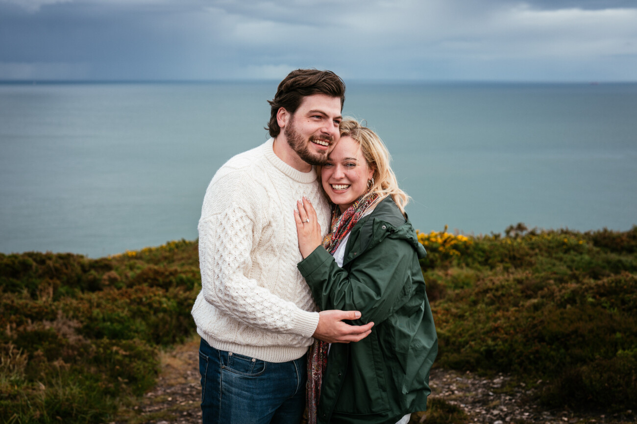 A smiling couple pose together outdoors on a grassy area with the sea and cloudy sky in the background. The woman shows off a ring on her hand.