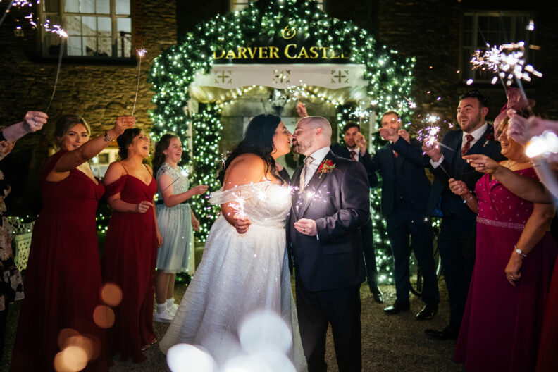Where to Actually Spend Your Wedding Budget (And What Your Guests Won't Remember) 8 A bride and groom kiss outside at night, surrounded by guests holding sparklers and festive lights, under a sign that reads "Darver Castle.