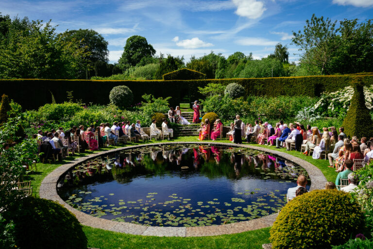 Best Wedding Photographers Ireland 7 Outdoor wedding ceremony by a circular pond, guests seated in rows around the water, surrounded by lush greenery under a blue sky.