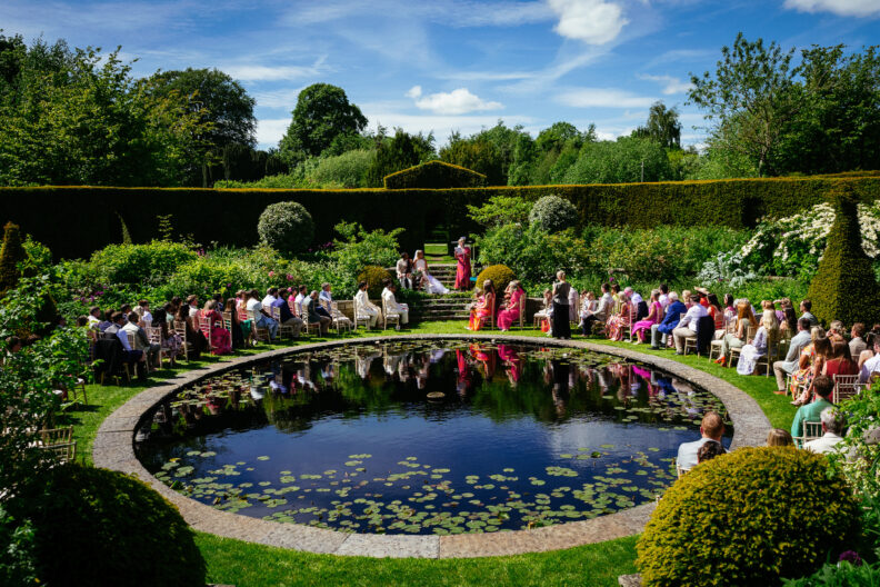 Best Wedding Photographers Ireland 8 Outdoor wedding ceremony by a circular pond, guests seated in rows around the water, surrounded by lush greenery under a blue sky.