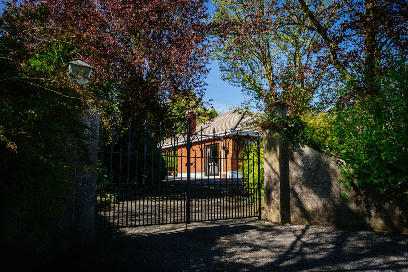 A black iron gate set in a stone wall opens to a drive leading to a red brick house, surrounded by trees and greenery under a clear blue sky—an idyllic setting reminiscent of Castle Durrow wedding photography.