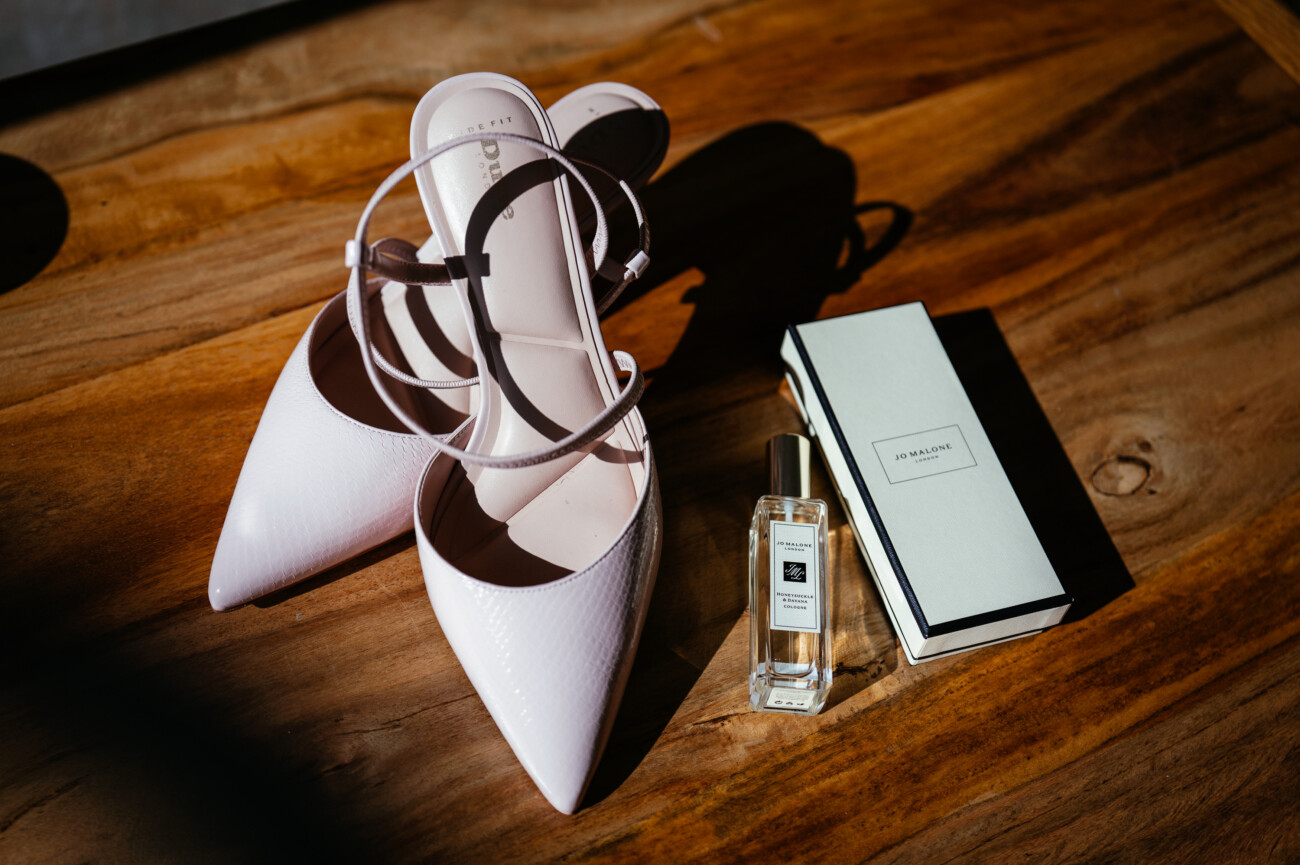 A pair of white heeled shoes, a bottle of Jo Malone perfume, and a Jo Malone box are placed on a wooden surface—capturing the timeless elegance often sought after in Castle Durrow Wedding Photography.