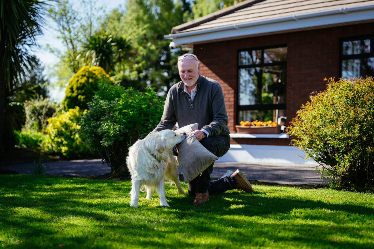 A man kneels on grass in a garden, smiling whilst stroking a white dog—a heartwarming moment reminiscent of the joyful candid shots often captured in Castle Durrow Wedding Photography. A brick house and various shrubs are visible in the background.