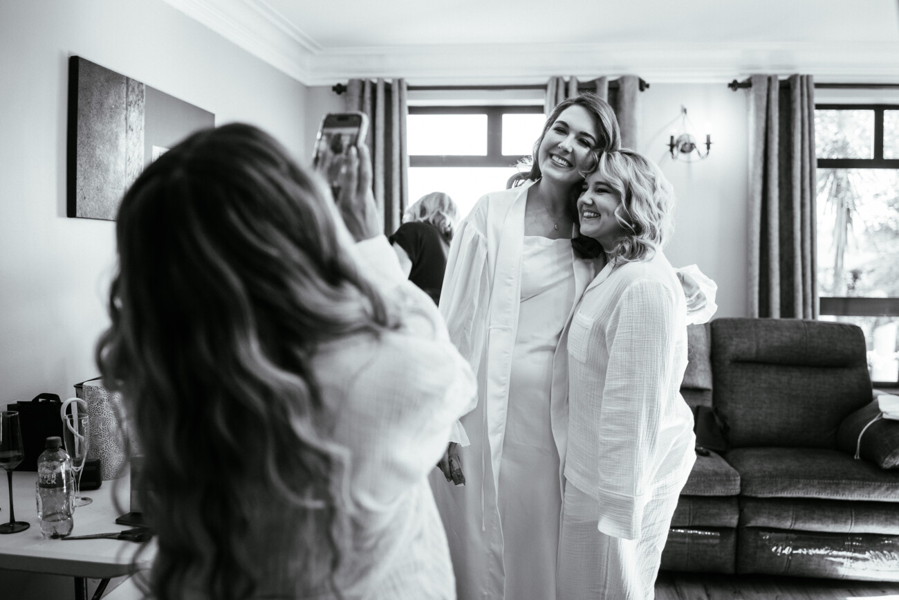 Two women in dressing gowns pose and smile for a photo being taken by another person in a sitting room with large windows and a sofa, capturing the joyful moments of a Castle Durrow Wedding Photography session.
