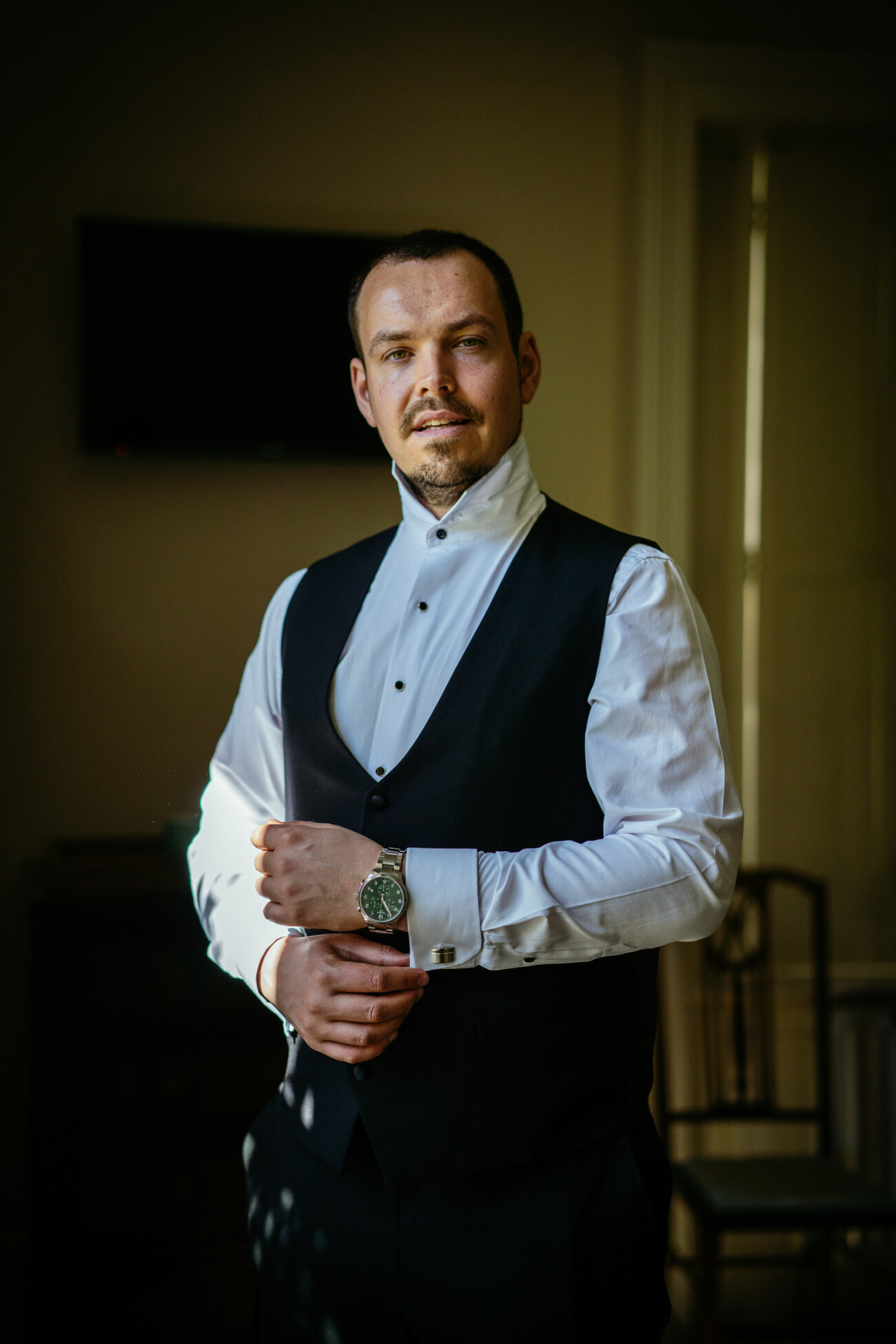 A man in a formal white shirt and black waistcoat stands indoors, adjusting his cufflink whilst looking at the camera—a timeless moment beautifully captured with Castle Durrow Wedding Photography.