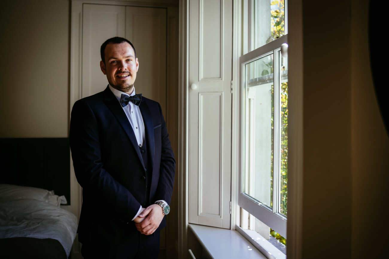 A man in a dinner jacket stands by a bright window in a bedroom at Castle Durrow, hands clasped, smiling at the camera—a perfect moment of Castle Durrow Wedding Photography.