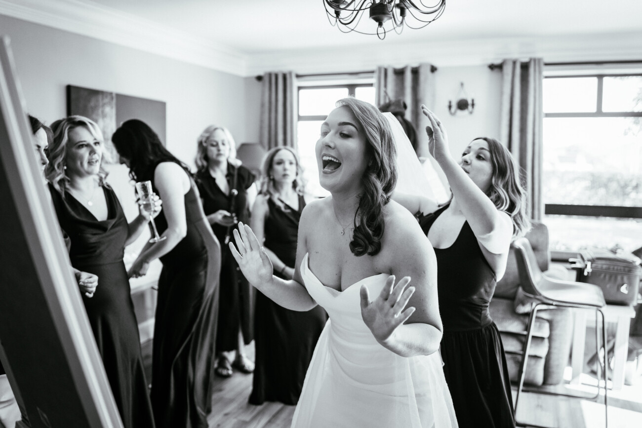 A bride in a strapless dress smiles with her hands raised as bridesmaids adjust her hair and gather round in a bright, sunlit room, perfectly capturing the joy and intimacy of Castle Durrow Wedding Photography.