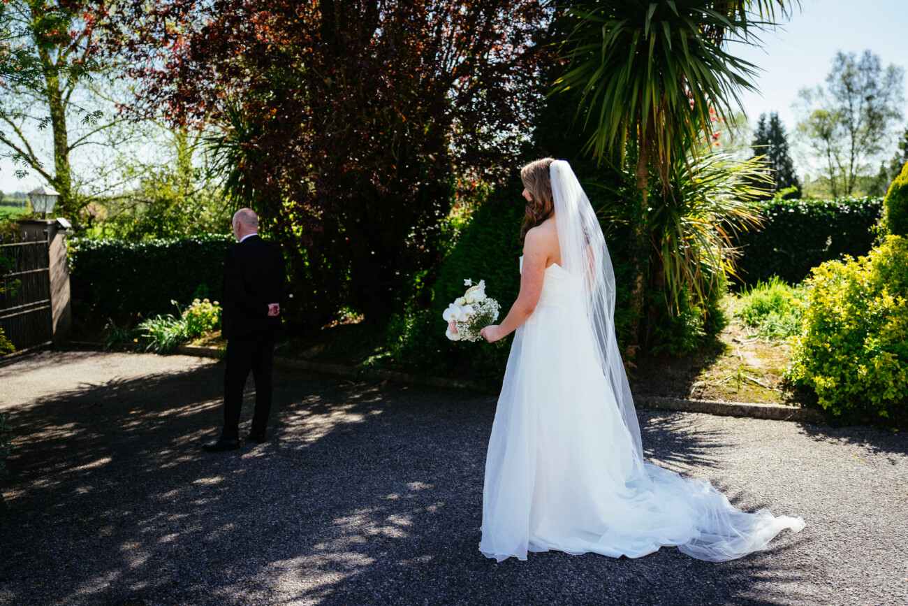 A bride in a white dress approaches a man in a suit standing with his back to her in a sunlit outdoor garden setting, capturing the timeless romance of Castle Durrow Wedding Photography.