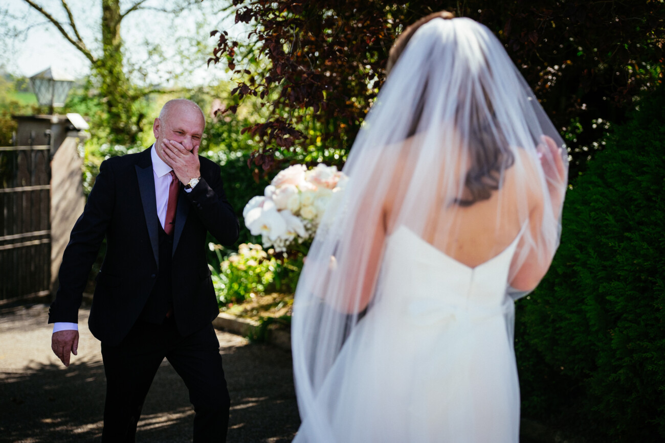 A man in a suit covers his mouth in surprise as he sees a bride in a white dress and veil outdoors on a sunny day, capturing the joy and emotion typical of Castle Durrow Wedding Photography.