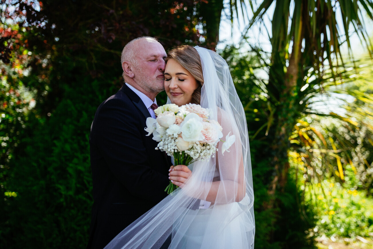 A man in a suit kisses a bride on the forehead as she holds a bouquet, both standing outdoors surrounded by greenery and sunlight, beautifully capturing the elegant charm of Castle Durrow Wedding Photography.