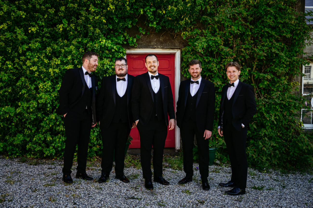Five men in black dinner jackets stand in front of a red door set in a green, leafy wall, posing and smiling for the camera—capturing the timeless elegance and joy found in Castle Durrow Wedding Photography.