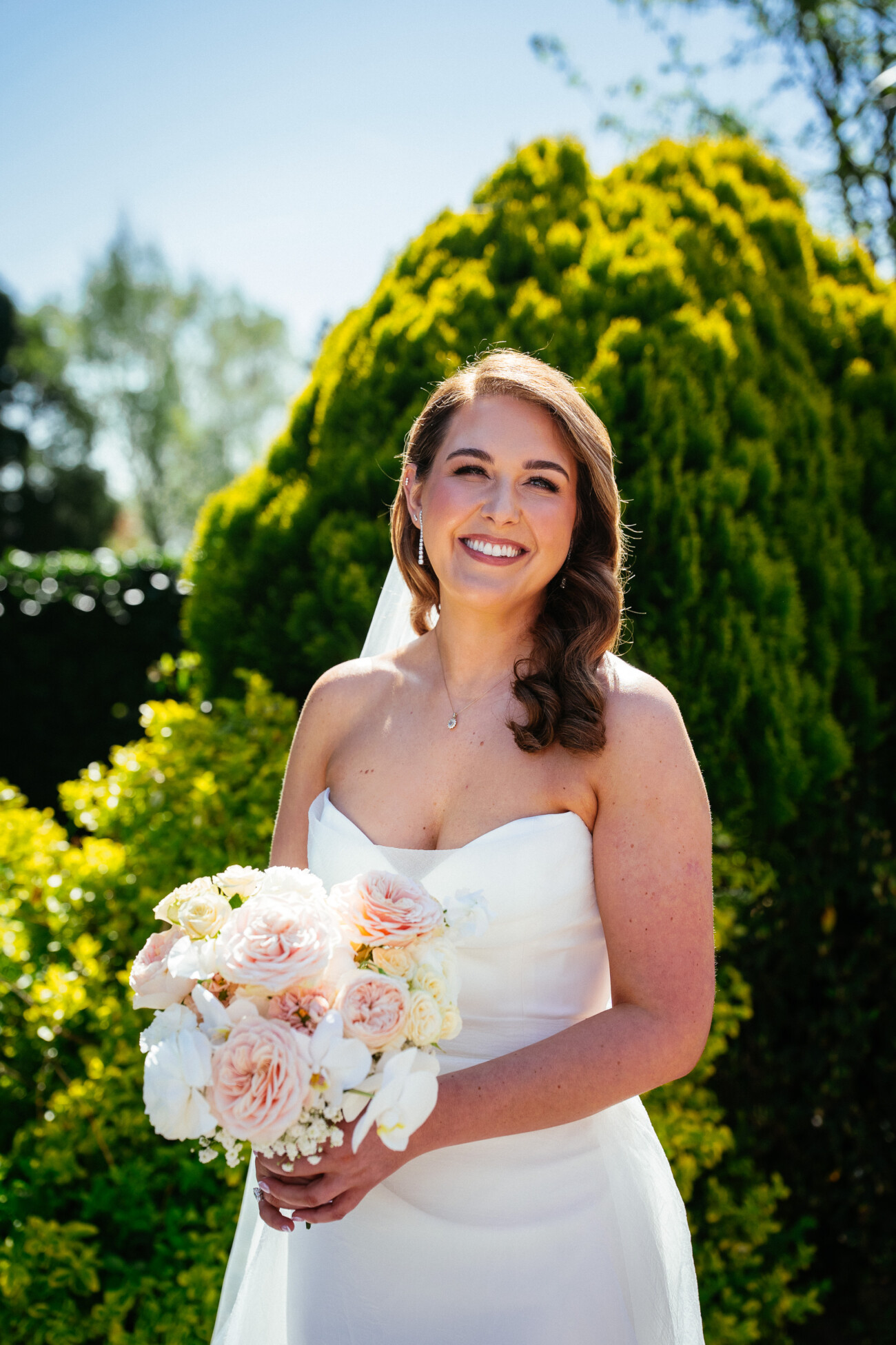 A bride in a strapless white dress holds a bouquet of pale flowers and smiles outdoors in front of green bushes on a sunny day, beautifully capturing the elegance and romance characteristic of Castle Durrow Wedding Photography.