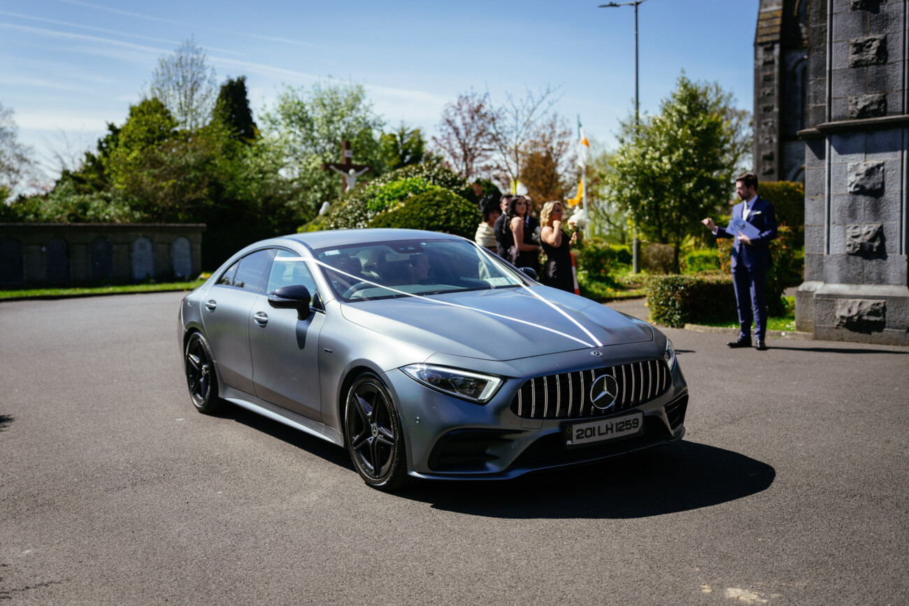 A silver Mercedes-Benz saloon is parked on a sunny street near stone buildings, with several people standing and talking in the background—a scene reminiscent of the elegance often captured in Castle Durrow Wedding Photography.