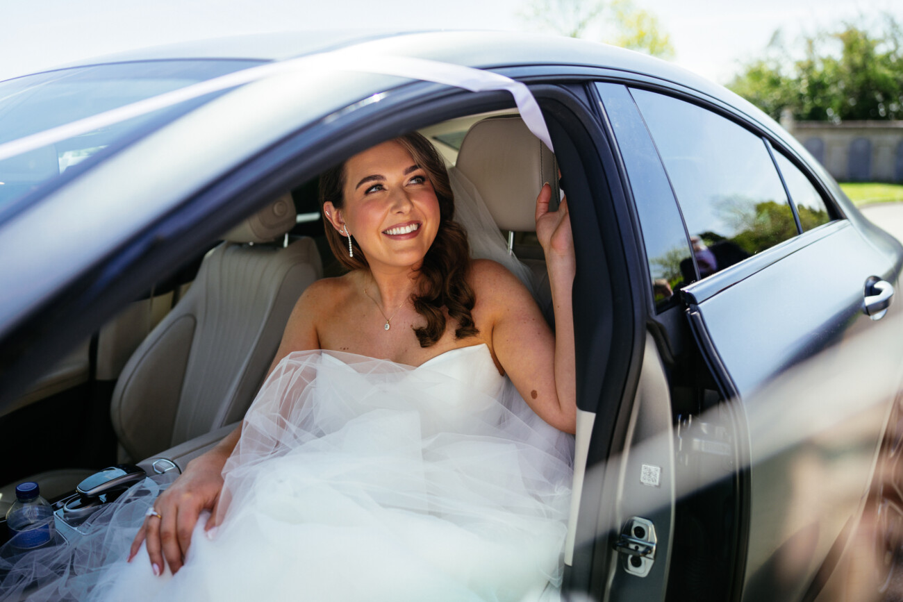 A woman in a wedding dress sits in the back seat of a car, smiling and holding a ribbon attached to the car door, with sunlight streaming in—a perfect moment captured by Castle Durrow Wedding Photography.