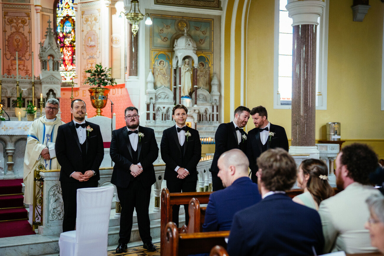 Five men in dinner jackets stand at the front of a church near the altar, facing the seated guests, with a priest in robes standing behind them—a classic moment beautifully captured through Castle Durrow Wedding Photography.