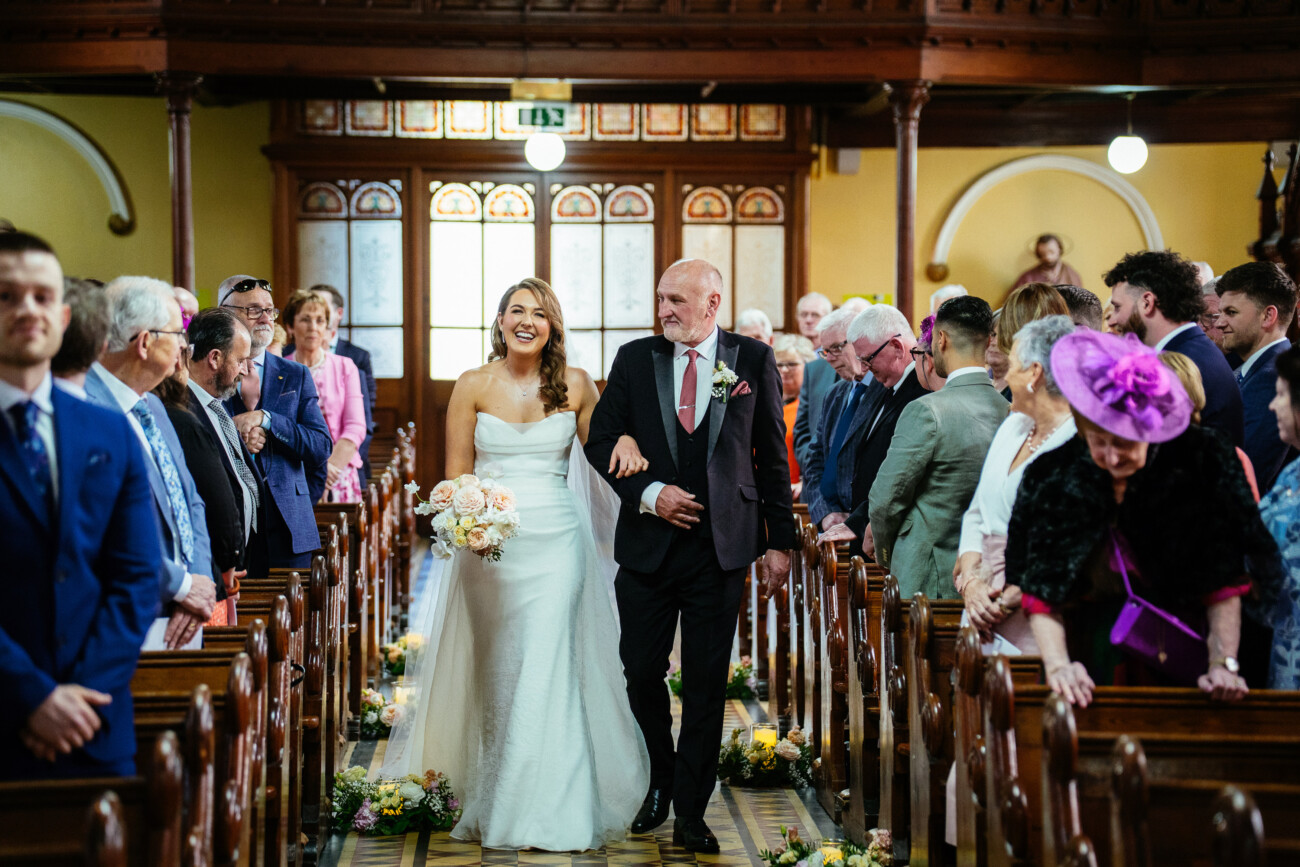 A bride in a white dress walks down the aisle with an older man, likely her father, as guests watch and smile inside a church—capturing the timeless elegance and emotion that defines Castle Durrow Wedding Photography.