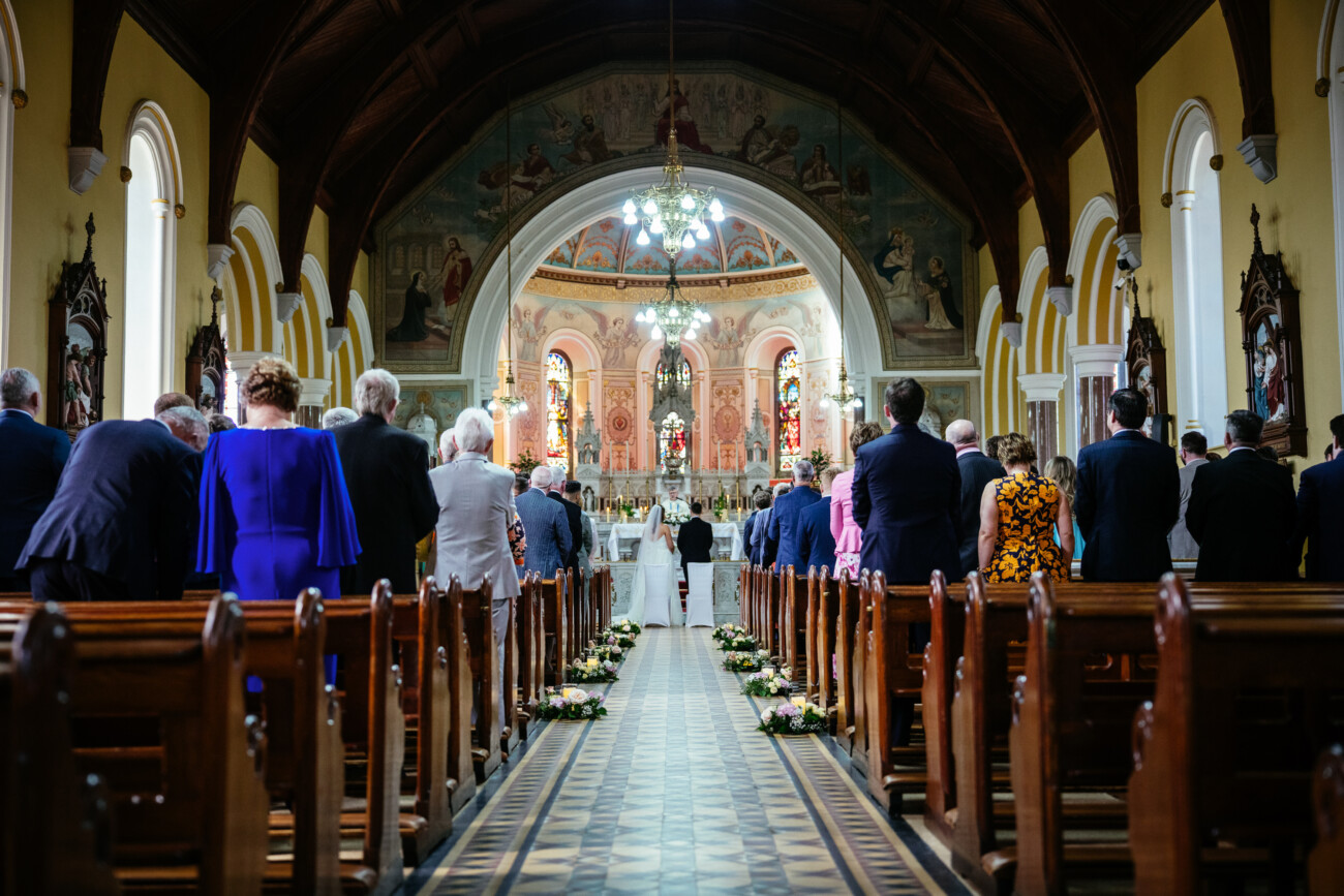 People are seated and standing in pews inside a beautifully decorated church, facing an altar at the front where a ceremony is taking place, capturing the timeless elegance often seen in Castle Durrow Wedding Photography.