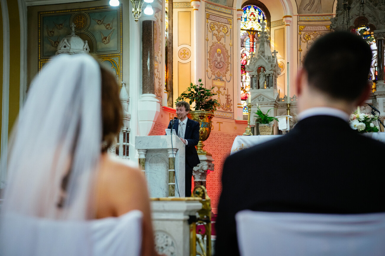 A man stands at a lectern speaking in a church, while a bride and groom sit in the foreground facing the altar—capturing an intimate moment reminiscent of timeless Castle Durrow Wedding Photography.