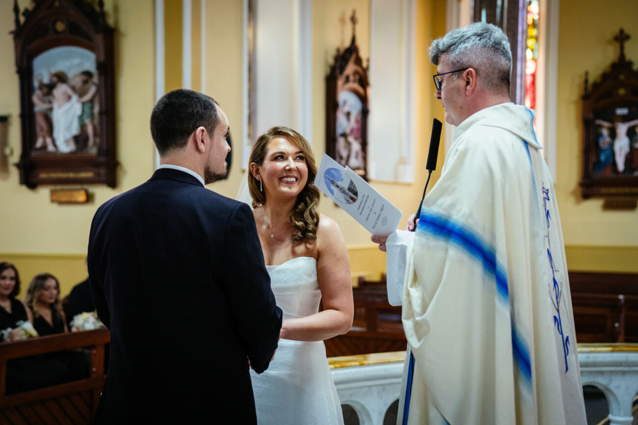 A bride and groom stand facing each other, holding hands, as a priest conducts a wedding ceremony inside a church—a timeless moment beautifully captured by Castle Durrow Wedding Photography.