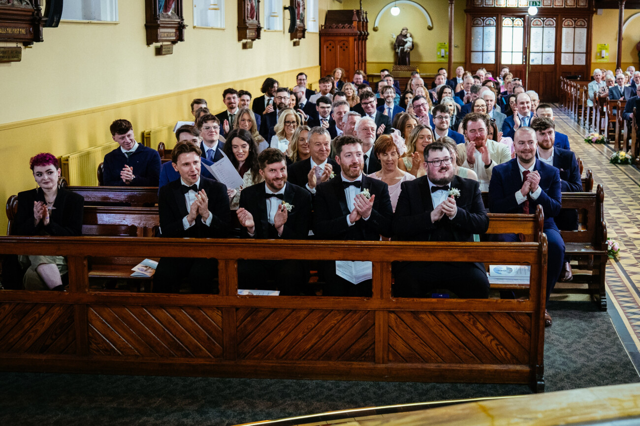 A group of people dressed formally sit in wooden pews inside a church, facing forwards and applauding, capturing a joyful moment through the lens of Castle Durrow Wedding Photography.