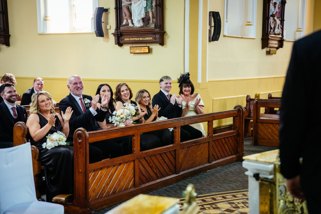 A group of people, some holding bouquets, sit in church pews and clap while facing someone standing at the front during a ceremony, beautifully captured in the timeless style of Castle Durrow Wedding Photography.