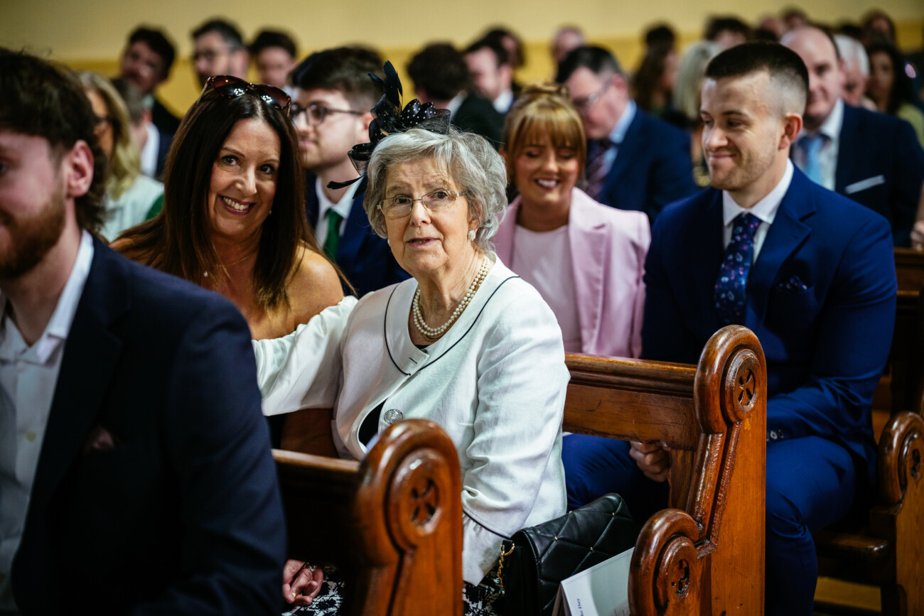 A group of people in formal attire sit on wooden pews at an indoor event, with a woman in white and pearls looking at the camera—a timeless moment beautifully captured by Castle Durrow Wedding Photography.