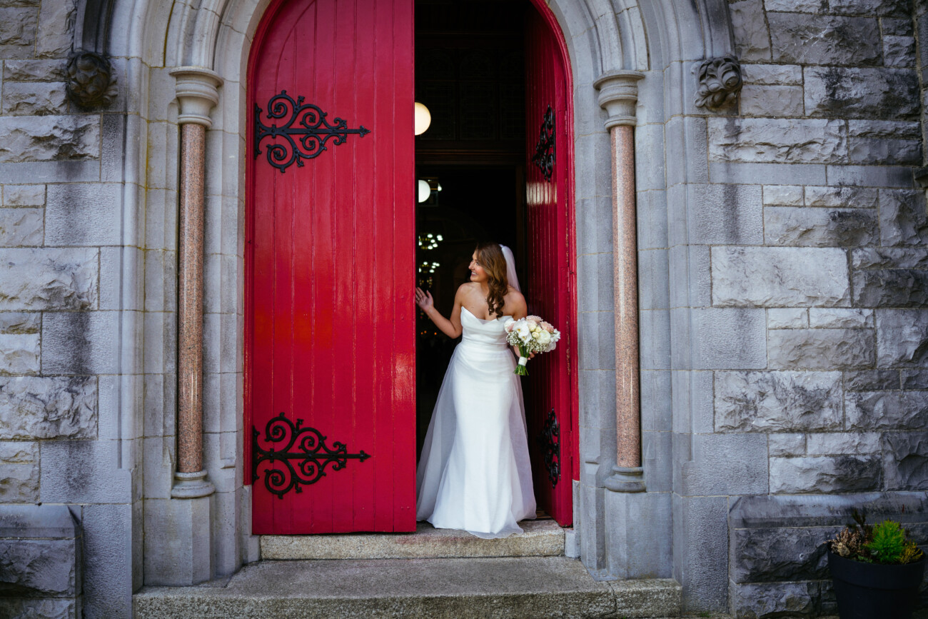 A bride in a white dress stands in the doorway of a stone building with large red doors, holding a bouquet of flowers—capturing the timeless elegance and romance that defines Castle Durrow wedding photography.