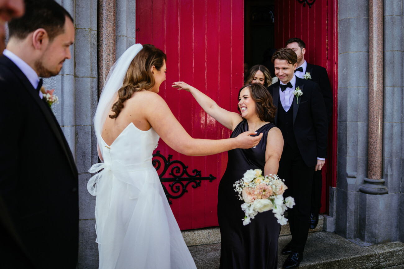 A bride in a white dress greets a smiling woman in a black dress holding flowers outside a building with red doors, whilst others stand behind them—capturing the joyful essence of Castle Durrow Wedding Photography.