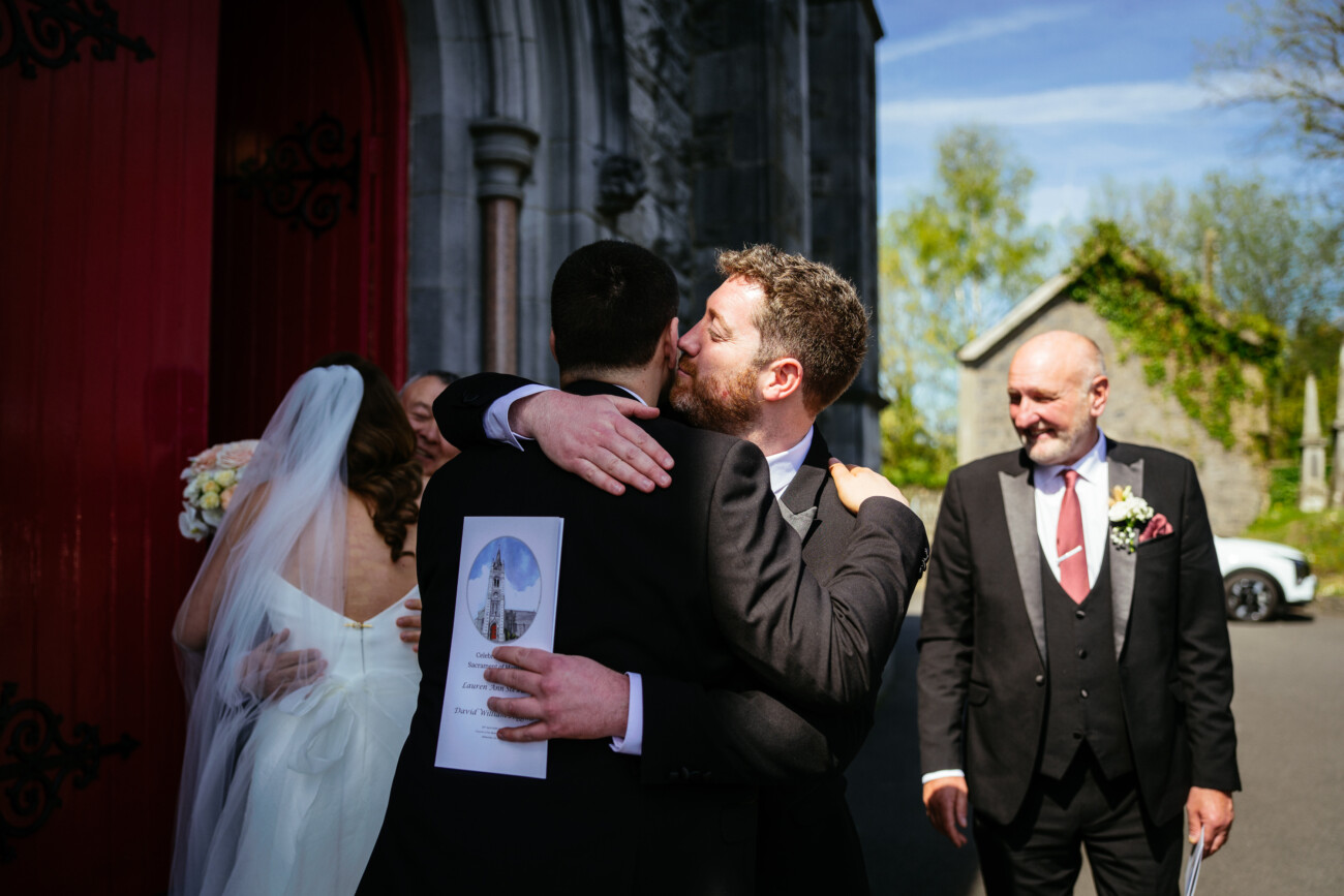 A man in a suit hugs another man at a wedding outside a church, while a bride and another man stand nearby in sunlight—capturing the warmth and emotion that define Castle Durrow Wedding Photography.