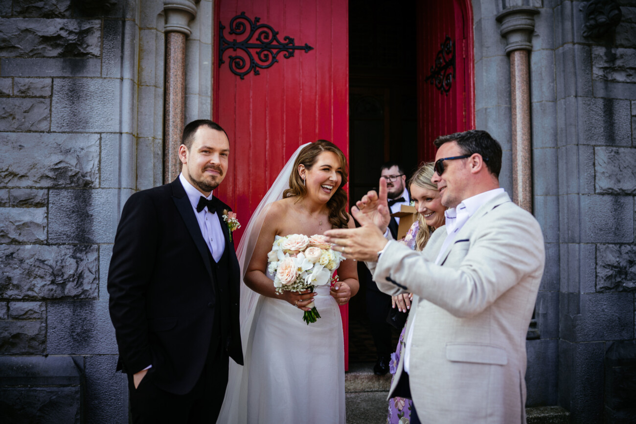 A bride and groom stand outside a church with red doors, smiling and talking with guests after their wedding ceremony, beautifully captured in the timeless style of Castle Durrow Wedding Photography.