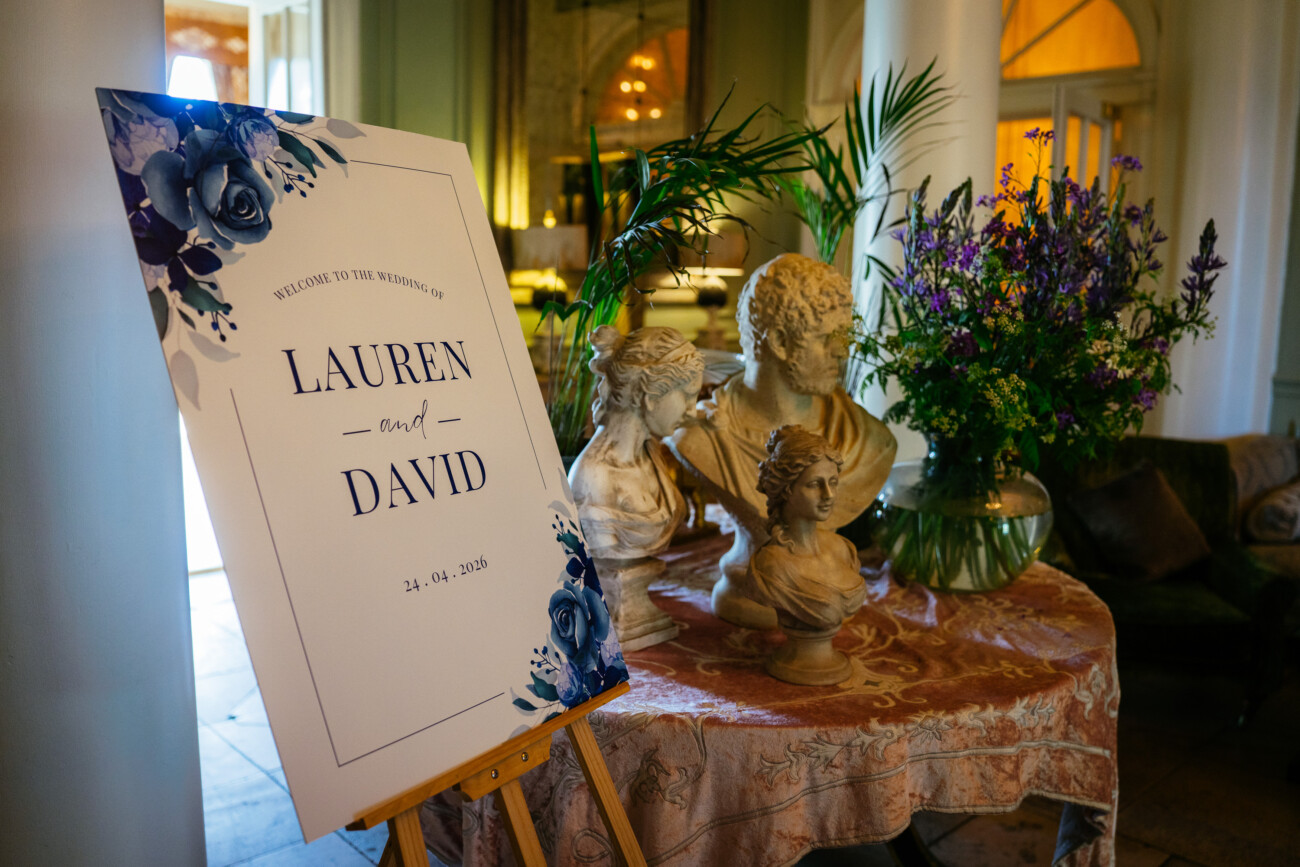 A wedding welcome sign for Lauren and David is displayed on an easel next to classical busts and a floral arrangement on a decorated table, beautifully captured through Castle Durrow Wedding Photography.