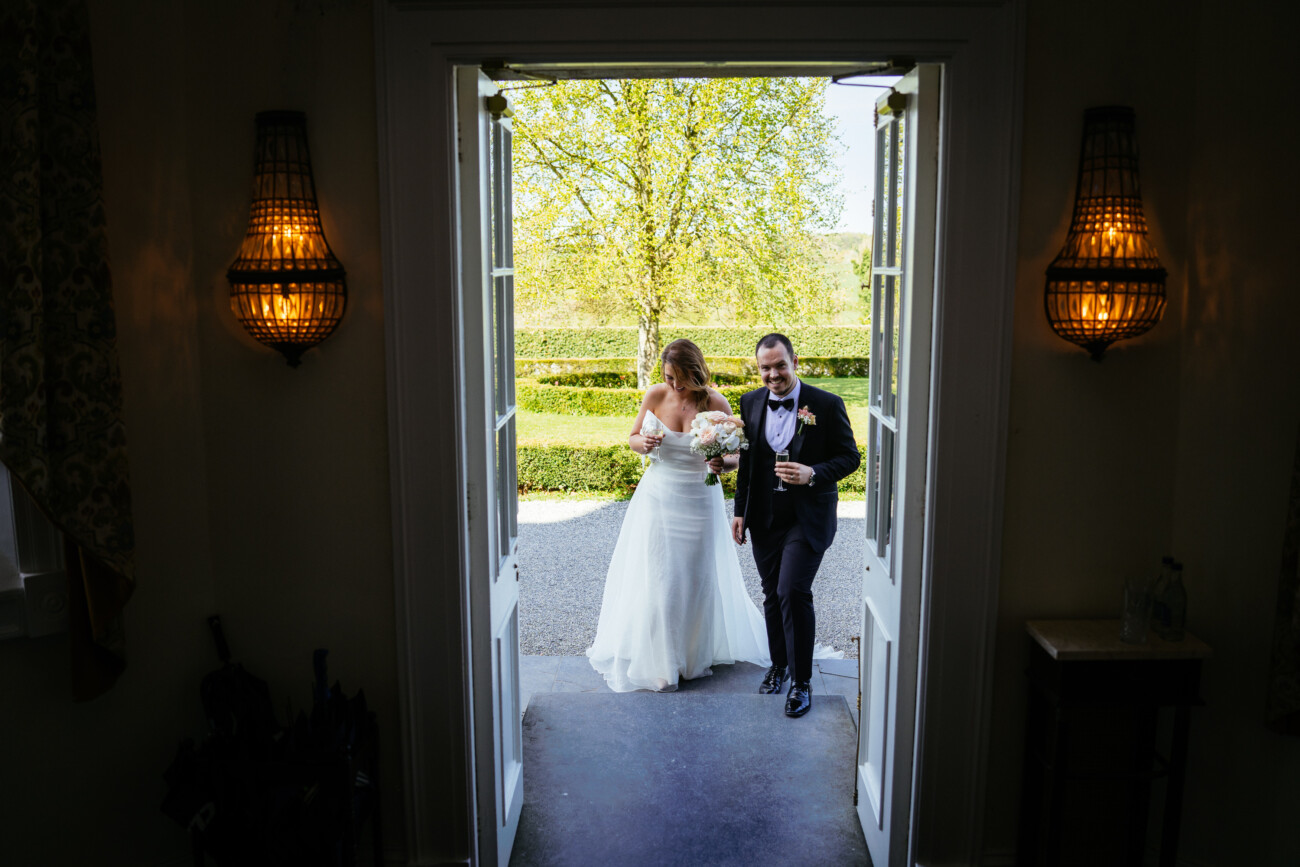 A bride and groom enter through open double doors, holding hands and smiling, with sunlight and greenery visible outside—capturing the timeless elegance of Castle Durrow Wedding Photography.