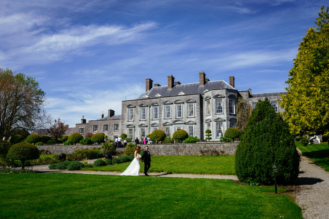 A bride and groom walk on a manicured lawn in front of a large stone mansion with gardens and people gathered outdoors under a blue sky, beautifully capturing the essence of Castle Durrow Wedding Photography.