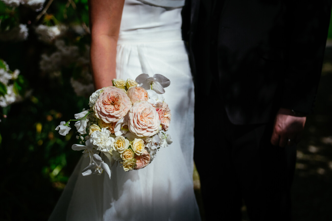 A bride in a white dress holds a bouquet of pale pink and cream flowers, standing next to someone in a black suit outdoors, beautifully capturing the elegance of Castle Durrow Wedding Photography.